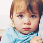 brown hair toddler with blue shirt and sad face held in parent's arms