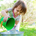 Toddler girl pours water from one bucket to another outside.
